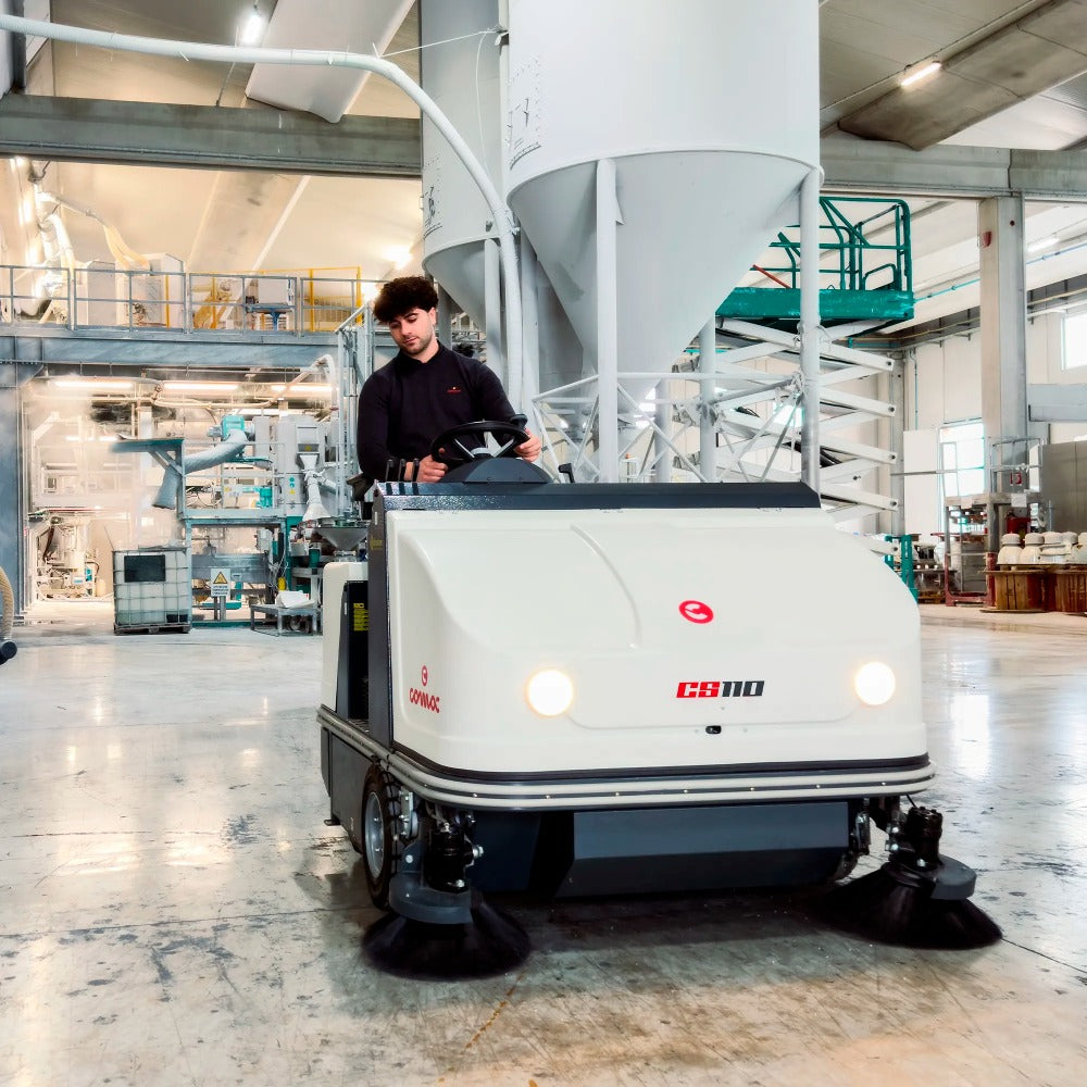 Person operating a Comac CS110B Ride On Sweeper cleaning machine in an industrial setting