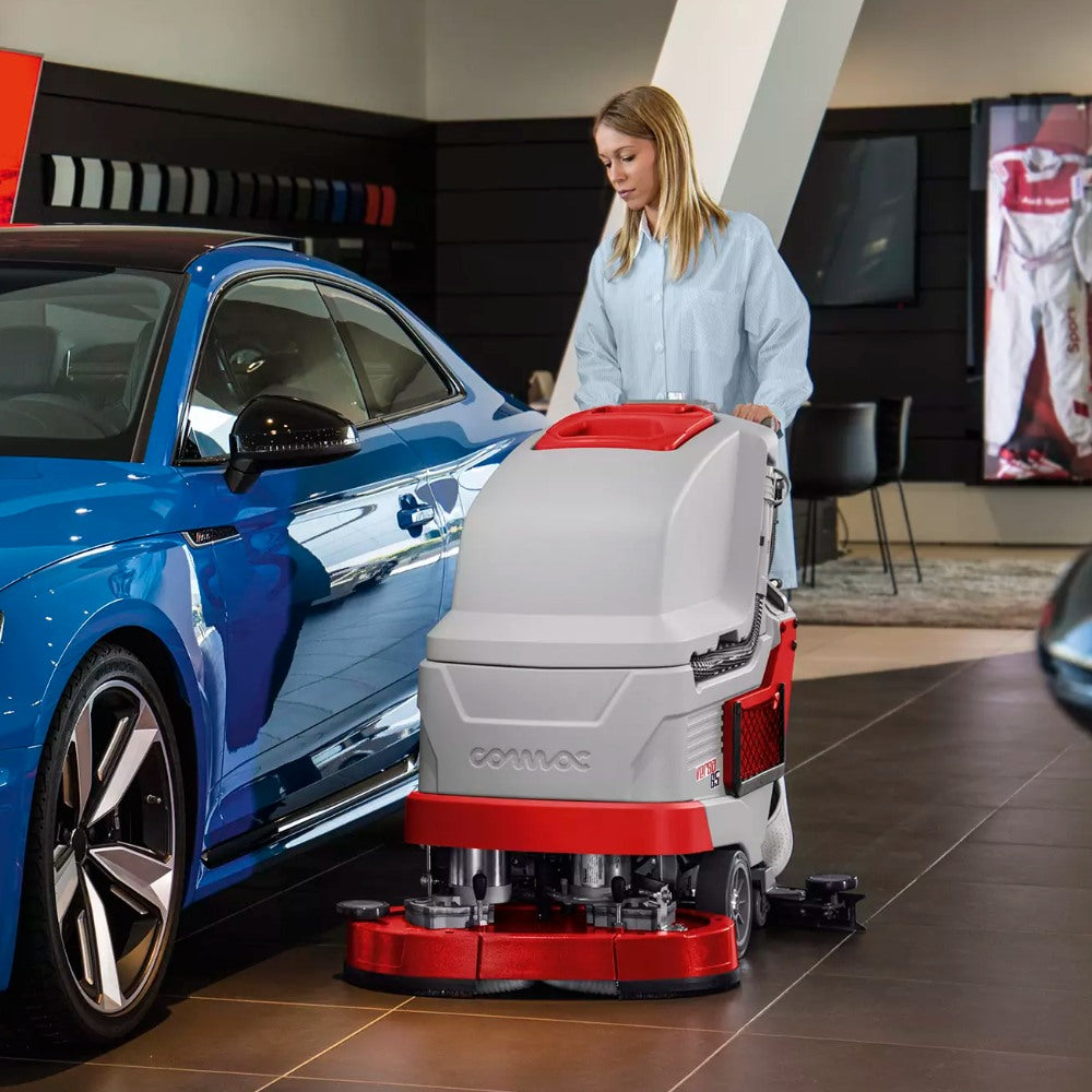 Person operating a floor cleaning machine next to a blue car in a showroom setting