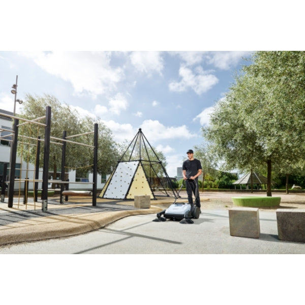 Person using a Nilfisk SW250 Walk Behind Sweeper in an outdoor setting with trees and playground equipment.