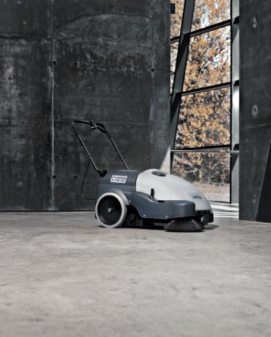 Nilfisk SW750 Walk-behind Sweeper on a concrete surface with a dark wall and trees in the background