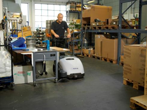 Man using a Nilfisk SW750 Walk-behind Sweeper in a warehouse setting with boxes and shelves.