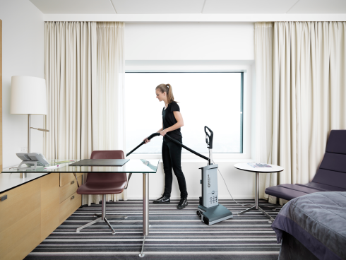 Person cleaning a room with a Nilfisk VU500 Upright Vacuum Cleaner 15" in a modern hotel room setting.