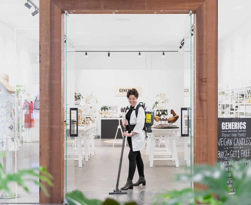 Person cleaning a store with a Pacvac Thrift 650 Backpack Vacuum Cleaner, surrounded by shelves and products.