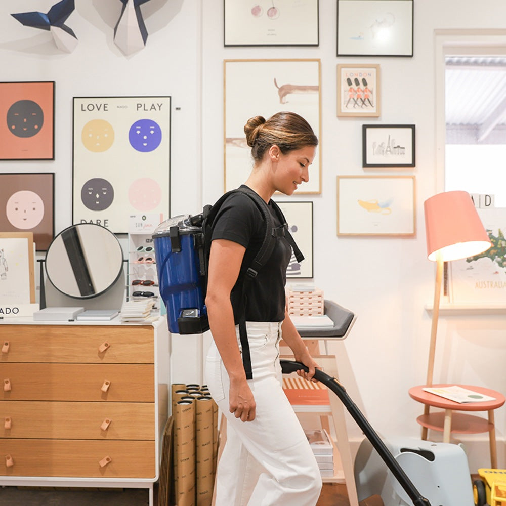 Woman using a backpack vacuum cleaner in a room with furniture and decor.