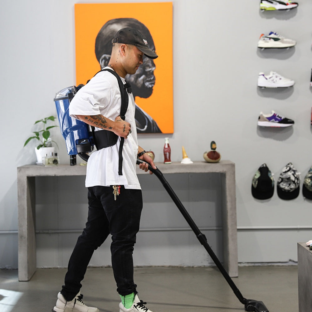 Person using a backpack vacuum cleaner in a room with shelves displaying shoes and hats.