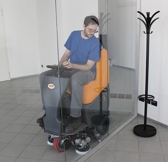 Person using a TSM Grande Brio Ride On 75-650 Scrubber in a tiled room with a coat rack.
