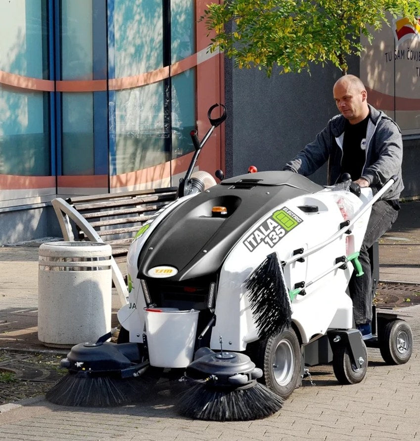 Person operating a large cleaning machine on a sidewalk with a building in the background