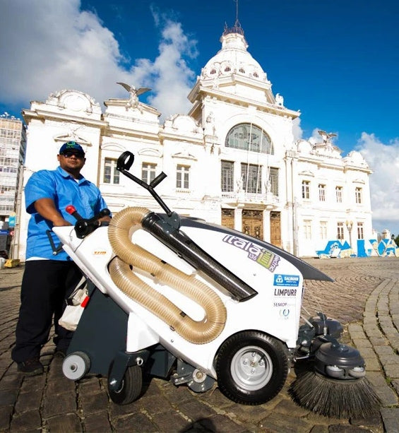 Person pushing a large cleaning machine in front of a white building with a blue sky.