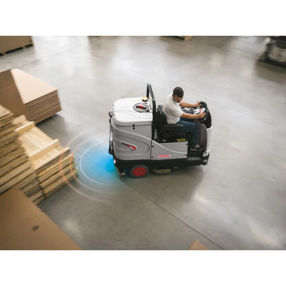Person operating a Comac C85 Essential Scrubber in an industrial setting with cardboard boxes.