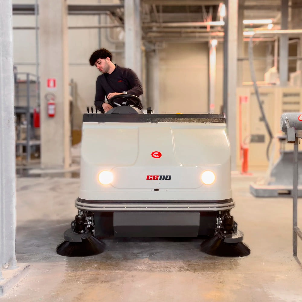 Person operating a Comac CS110B Ride On Sweeper in an industrial setting