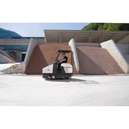 Person operating a Comac CS120B Ride On Sweeper on a concrete surface with a building and mountains in the background