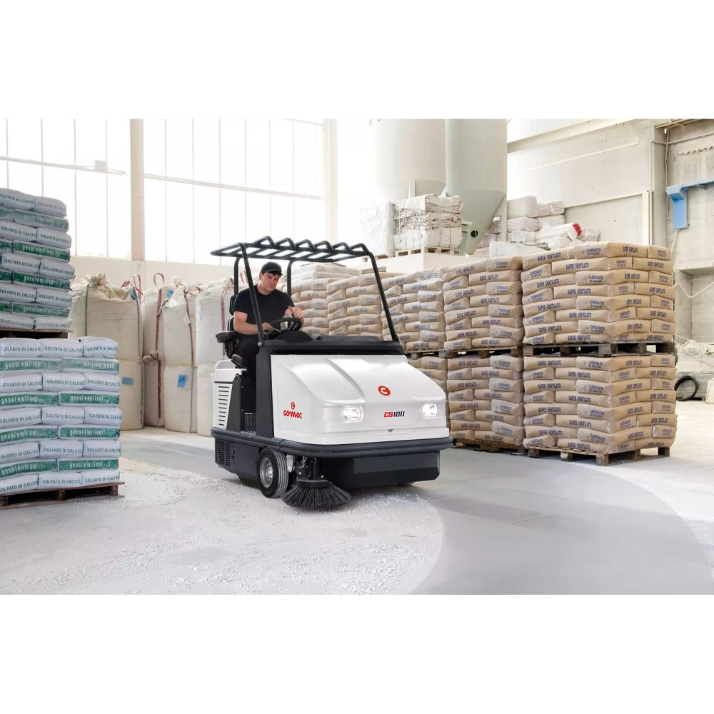 Person operating a Comac CS120B Ride On Sweeper in a warehouse setting with stacked pallets of goods