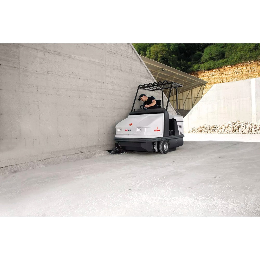 Person operating a Comac CS120B Ride On Sweeper on a concrete surface with a concrete wall and greenery in the background