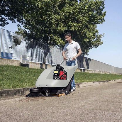 Person operating a floor sweeping machine on a road with trees and a fence in the background