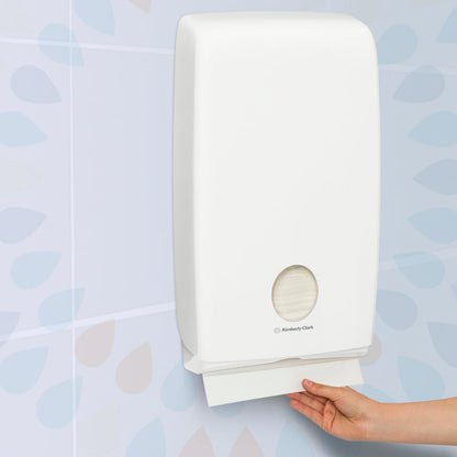 White paper towel dispenser on a tiled wall with a hand reaching for a roll of paper towels.
