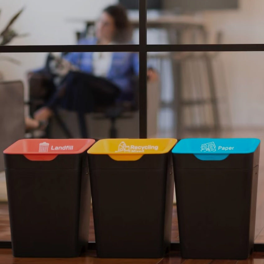 Three recycling bins labeled 'Landfill', 'Recycling', and 'Paper' with a blurred office background.