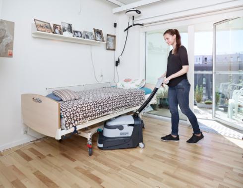 Person using a Nilfisk SC250 Scrubber on a wooden floor in a room with a bed and large window.