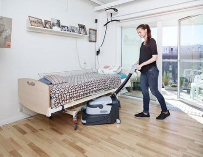 Person using a Nilfisk SC250 Scrubber on a wooden floor in a room with a bed and large window.
