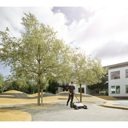 Person using a lawn mower in a park with trees and buildings in the background