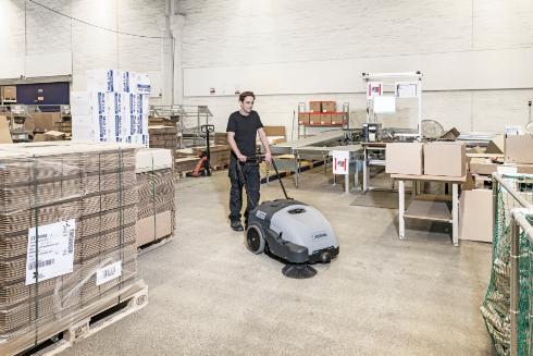Person operating a Nilfisk SW750 Walk-behind Sweeper cleaning machine in a warehouse setting