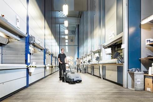 Person using a Nilfisk SW750 Walk-behind Sweeper cleaning machine in a modern warehouse setting