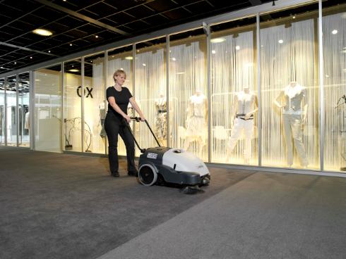 Person using a Nilfisk SW750 Walk-behind Sweeper cleaning machine in a modern indoor setting with glass walls.