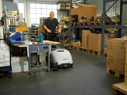Man using a Nilfisk SW750 Walk-behind Sweeper in a warehouse setting with boxes and shelves.