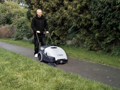 Man operating a Nilfisk SW750 Walk-behind Sweeper on a grassy area with trees in the background