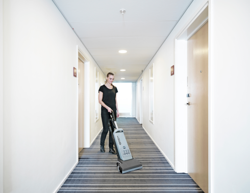 Person using a Nilfisk VU500 Upright Vacuum Cleaner 15" in a hallway with white walls and striped carpet.