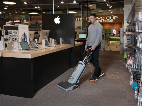 Person using a Nilfisk VU500 Upright Vacuum Cleaner in a store setting with laptops on display.