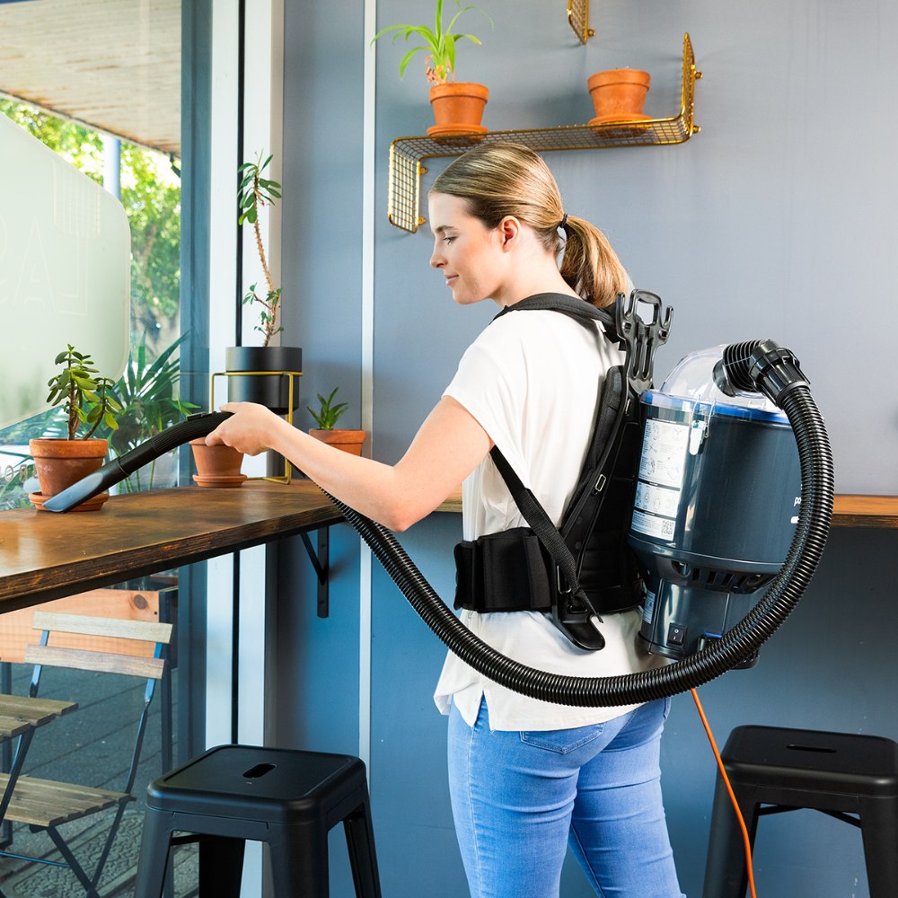 Person using a backpack vacuum cleaner in an indoor setting with plants and furniture