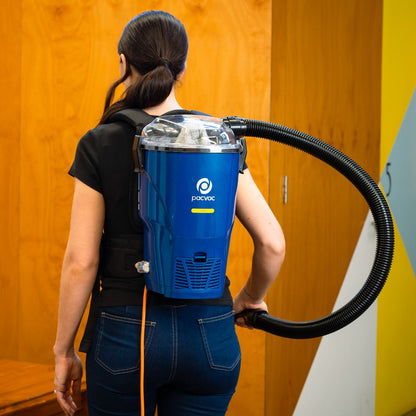 Person wearing a blue backpack vacuum cleaner with a long hose against a wooden wall.