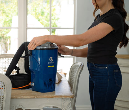 Person using a blue vacuum cleaner in a room with large windows.