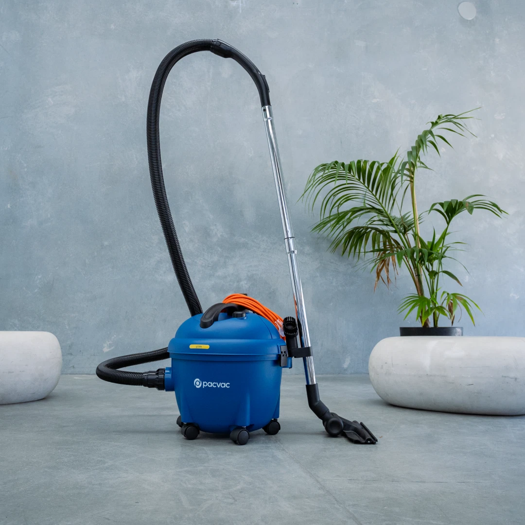 Blue handheld Pacvac Glide 300 Barrel Vacuum Cleaner with a long black hose on a gray surface with a plant in the background.