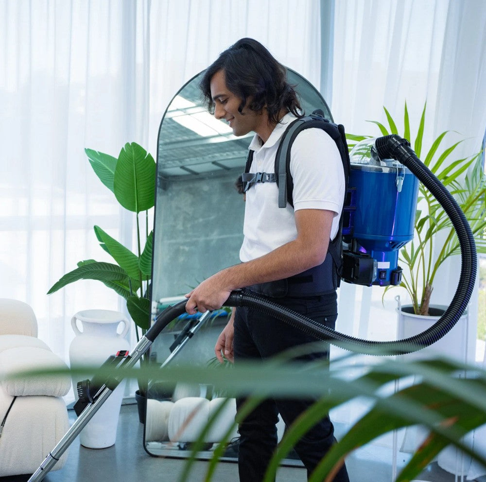 Person using a Pacvac Superpro vacuum cleaner in a room with plants and furniture.