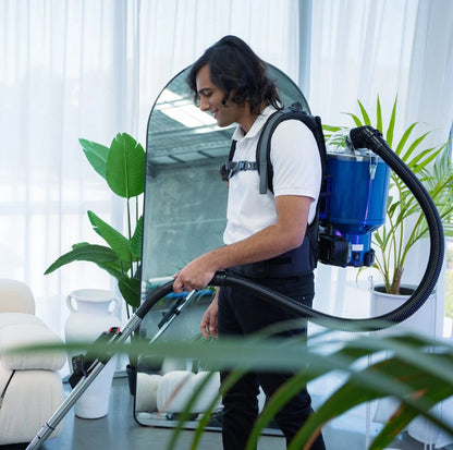 Person using a Pacvac Superpro vacuum cleaner in a room with plants and furniture.