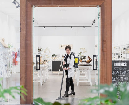 Person cleaning a store with a Pacvac Thrift 650 Backpack Vacuum Cleaner, surrounded by shelves and products.
