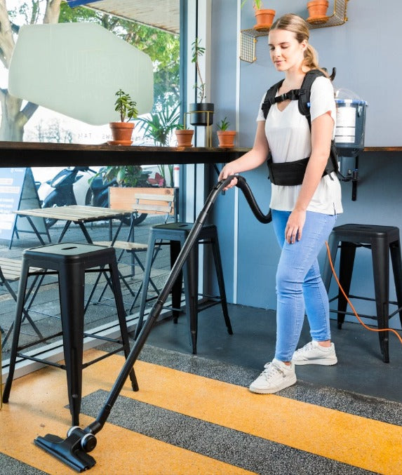 Person using a Pacvac Thrift 650 Backpack Vacuum Cleaner in an cafe setting with tables and chairs.