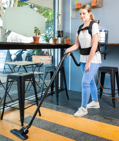 Person using a Pacvac Thrift 650 Backpack Vacuum Cleaner in an cafe setting with tables and chairs.