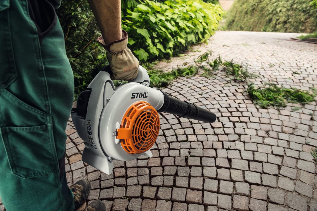 Person using a STIHL leaf blower on a paved path with greenery.
