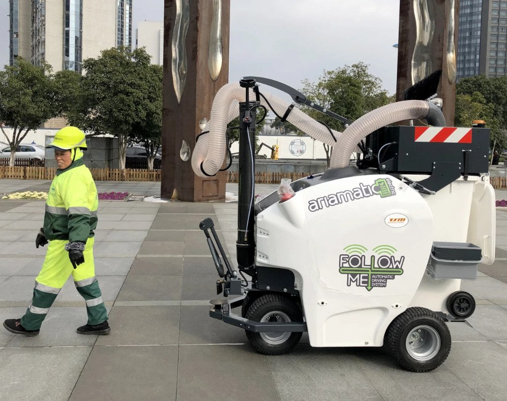 Worker in high-visibility clothing operating a self-propelled TSM Ariamatic 240 cleaning machine on a tiled pavement.
