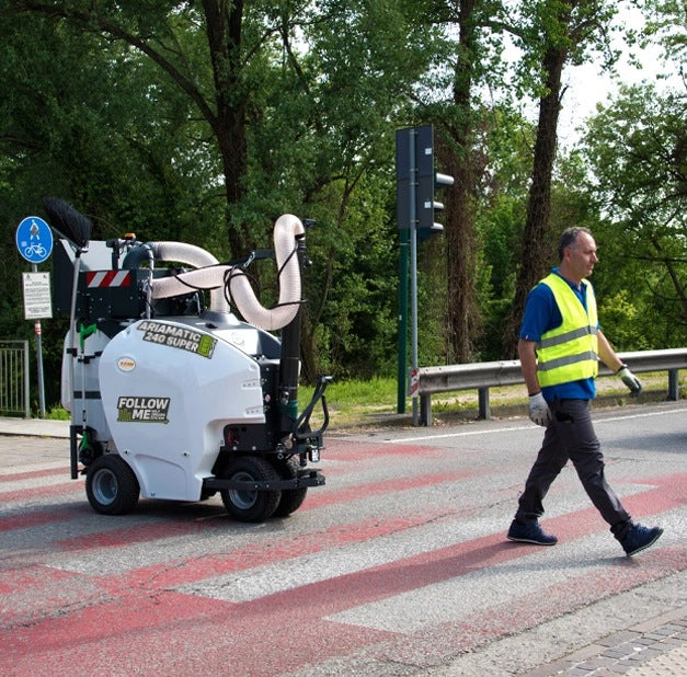 Person in high-visibility clothing walking next to a self-driving TSM Ariamatic 240 on a road.