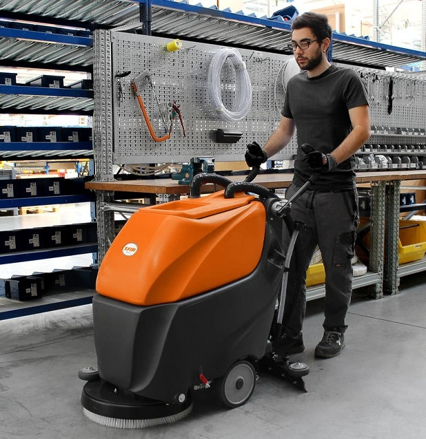 Person standing next to an orange and black TSM Grande Brio 50B Scrubber Dryer machine in a workshop setting.