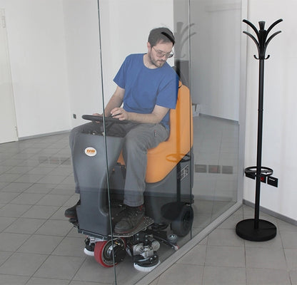 Person using a TSM Grande Brio Ride On 75-650 Scrubber in a tiled room with a coat rack.