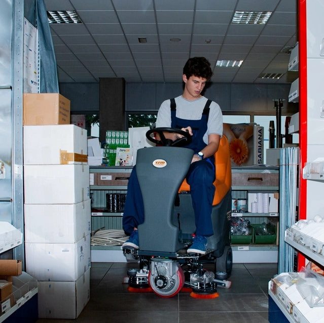 Person sitting on a TSM Grande Brio Ride On 75-650 Scrubber cleaning machine in a warehouse setting