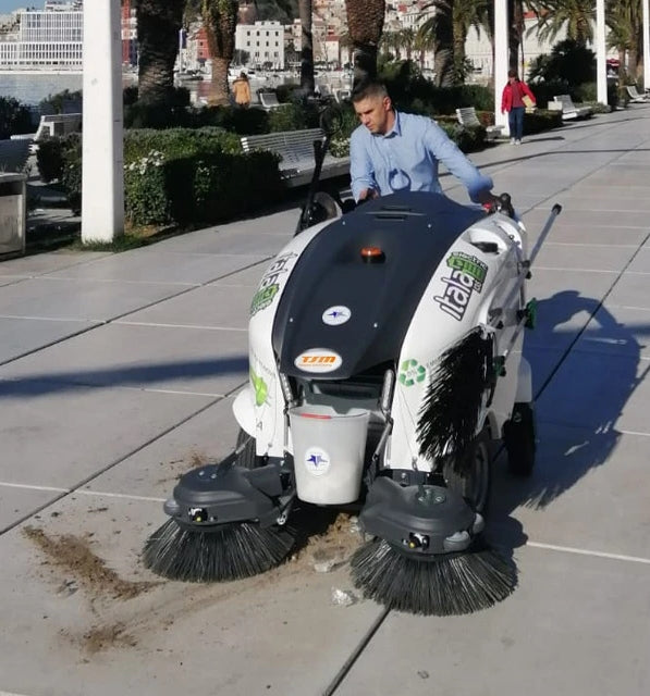 Person operating a street cleaning machine on a sidewalk with a cityscape in the background