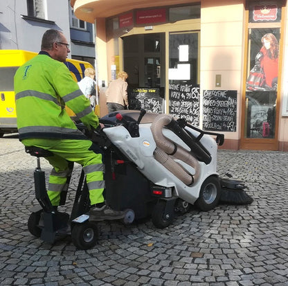 Person using a TSM Itala 135 BT Sidewalk Sweeper on a cobblestone street with a store in the background.