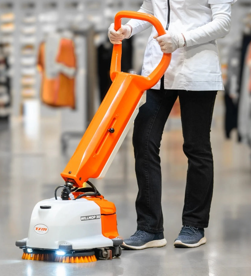Person using an orange and white TSM Willmop 35 Scrubber Dryer machine in a blurred indoor setting